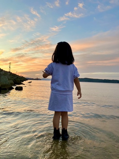 海に足をつけて夕日を見ている少女の後ろ姿