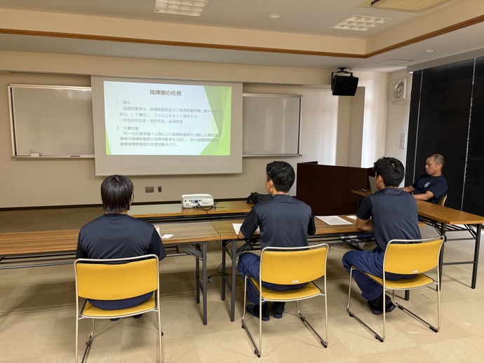 初任教育中の学生が、指揮調査隊員に指揮調査隊の役割をプロジェクターを使用し、スライドを投影しながら教えてもらっている写真。
