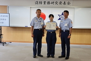 写真：林 副士長が西本消防局長と上辻中消防署長にはさまれ、賞状を持って立っている。