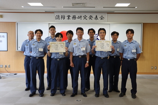 写真：西本消防局長を中心に発表者全員（9名）が立っている。